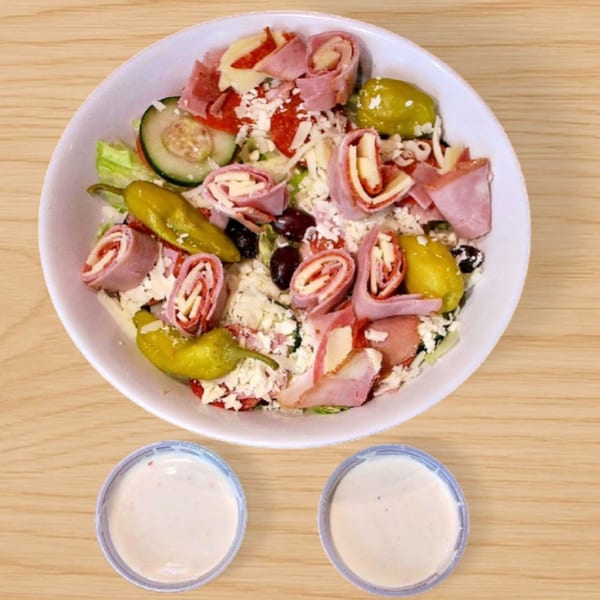 A Babe's Pizza salad with ham, cheese, pepperoni, olives, and peppers, with two dressing cups.