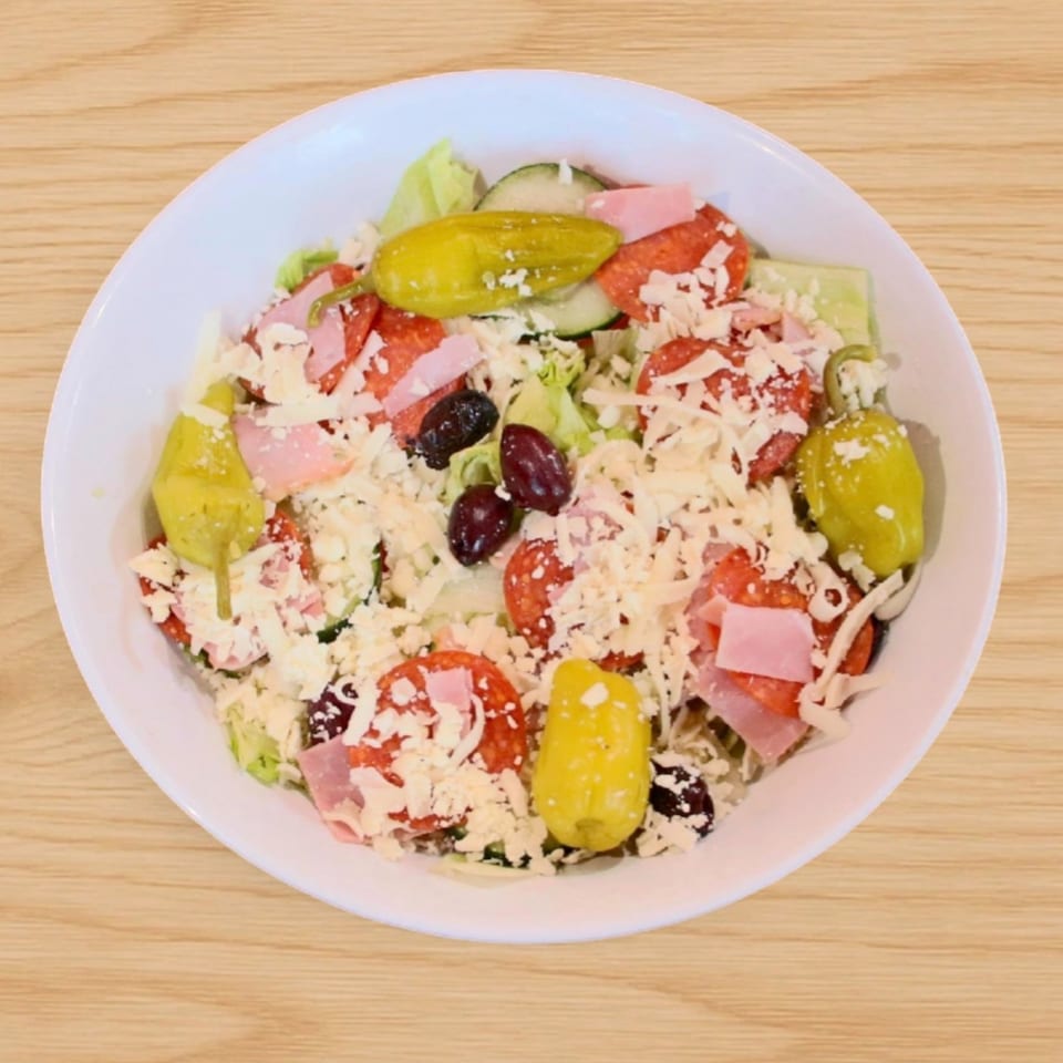 A white bowl of Babe's Pizza salad with pepperoni, ham, olives, pepperoncini, and shredded cheese.