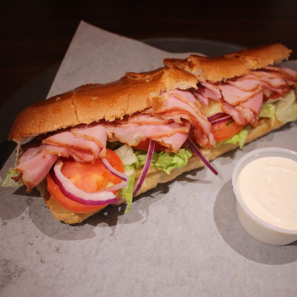A Babe's Pizza sub sandwich with ham, lettuce, tomatoes, and red onions, with a side of sauce.