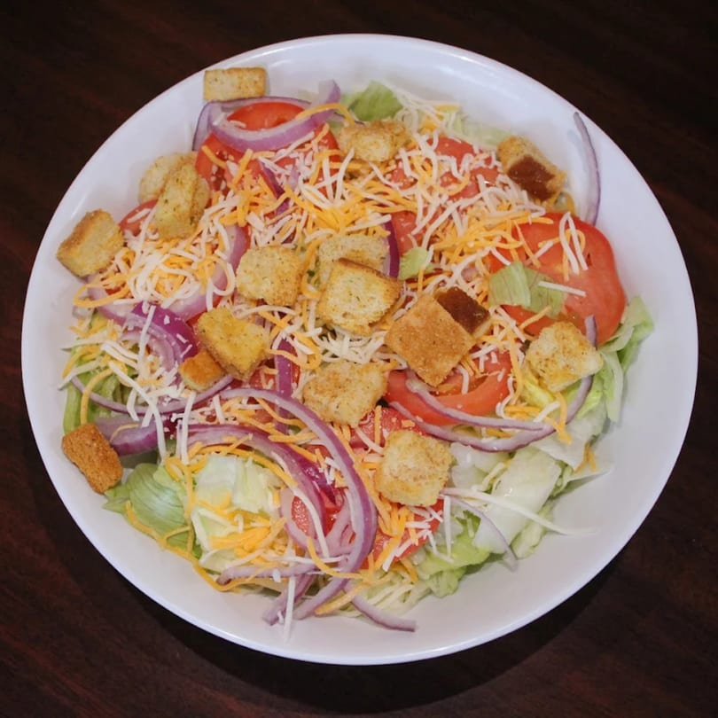 A fresh side salad with lettuce, tomatoes, red onions, shredded cheese, and croutons in a white bowl.