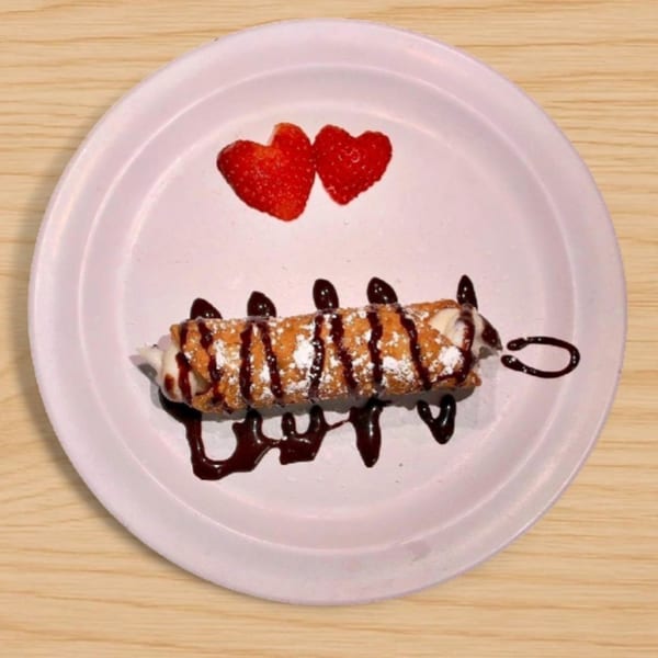 A cannoli drizzled with chocolate and powdered sugar, served with two heart-shaped strawberries on a white plate.